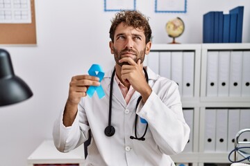 Young hispanic doctor man holding blue ribbon serious face thinking about question with hand on...