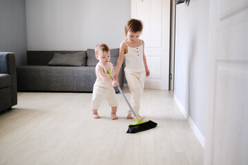 Children siblings with mop sweep floor in bright living room, help with housework and sibling...