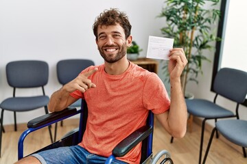 Young handsome man holding covid record card sitting on wheelchair smiling happy pointing with hand...