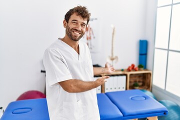 Young handsome physiotherapist man working at pain recovery clinic inviting to enter smiling...