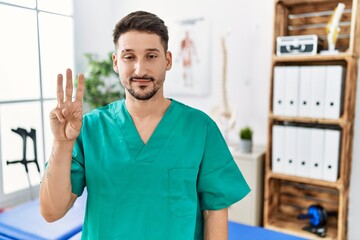 Young physiotherapist man working at pain recovery clinic showing and pointing up with fingers number three while smiling confident and happy.