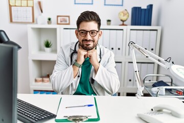 Young man with beard wearing doctor uniform and stethoscope at the clinic laughing nervous and excited with hands on chin looking to the side