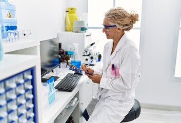 Middle age blonde woman wearing scientist uniform writing on clipboard working at laboratory