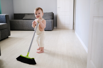 Toddler 1 year old with broom sweeps floor in bright room in Scandinavian style, montessori development, helping his mother and independence