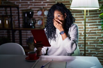 Middle age hispanic woman using touchpad sitting on the table at night shocked covering mouth with...