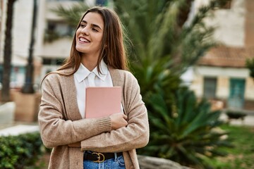 Young hispanic student girl smiling happy holding book at the city.