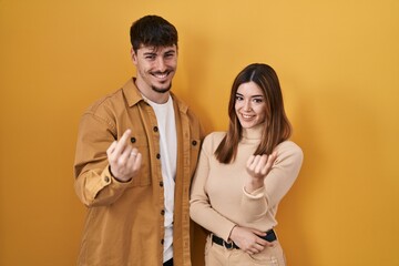 Young hispanic couple standing over yellow background beckoning come here gesture with hand inviting welcoming happy and smiling