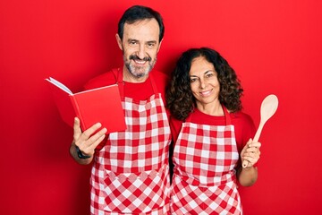 Middle age couple of hispanic woman and man wearing professional apron reading cooking recipe book smiling with a happy and cool smile on face. showing teeth.
