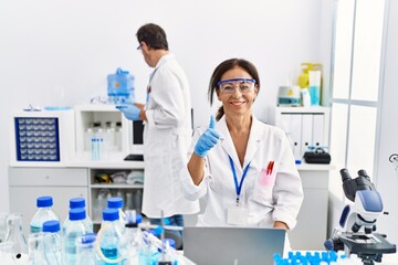 Middle age woman working at scientist laboratory smiling happy and positive, thumb up doing excellent and approval sign