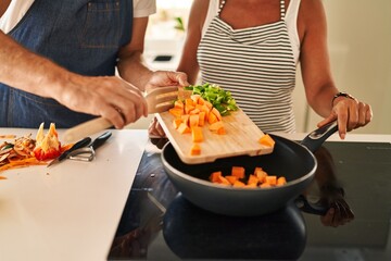 Middle age hispanic couple pouring food on frying pan at kitchen