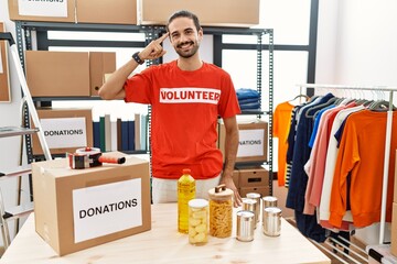 Young hispanic man wearing volunteer t shirt at donations stand smiling pointing to head with one...