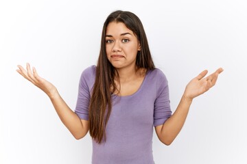 Young brunette woman standing by isolated background clueless and confused expression with arms and...