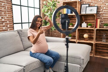 Young pregnant woman recording vlog tutorial with smartphone at home smiling making frame with...