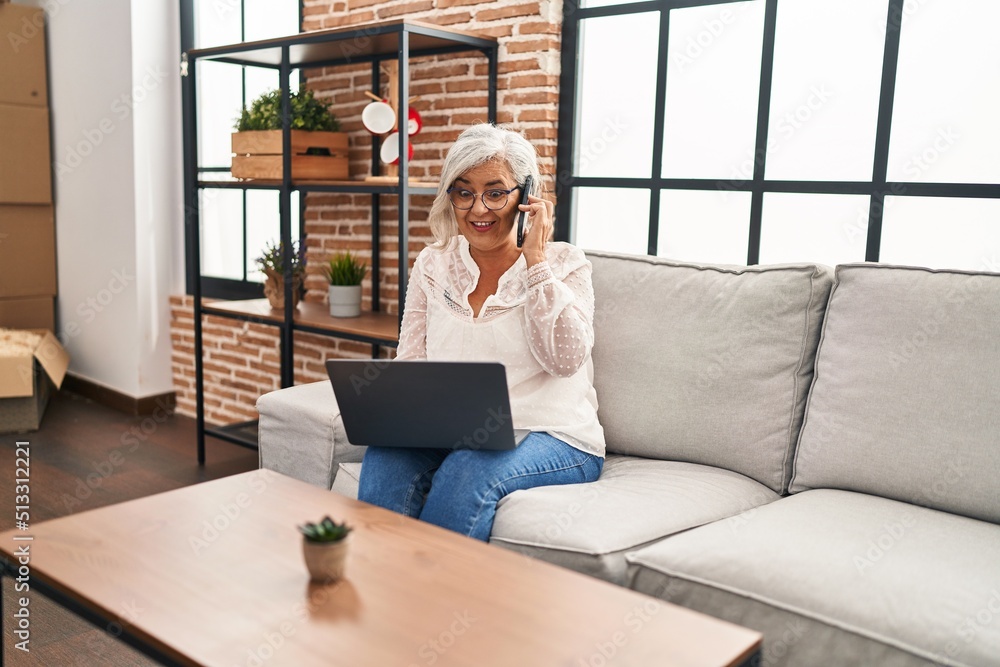 Poster Middle age woman talking on the smartphone using laptop at home