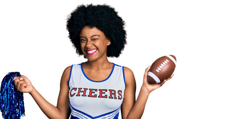 Young african american woman wearing cheerleader uniform holding pompom and football ball winking...