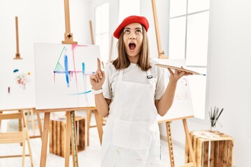 Young artist woman painting on a canvas at art studio amazed and surprised looking up and pointing with fingers and raised arms.