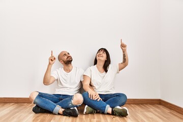 Young hispanic couple smiling happy pointing with finger to the ceiling at empty new home.