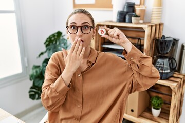 Young blonde woman holding virtual currency bitcoin at the office covering mouth with hand, shocked...