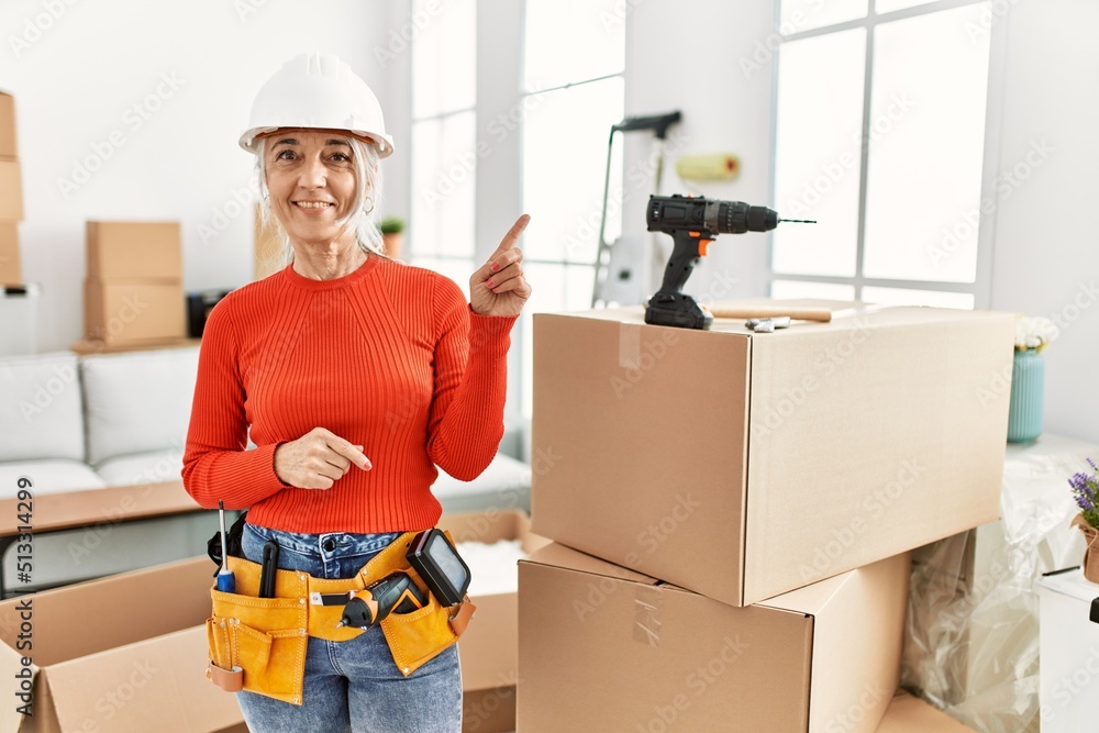 Canvas Prints Middle age grey-haired woman wearing hardhat standing at new home with a big smile on face, pointing with hand and finger to the side looking at the camera.