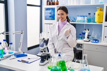 Young brunette woman working at scientist laboratory smiling happy and positive, thumb up doing...
