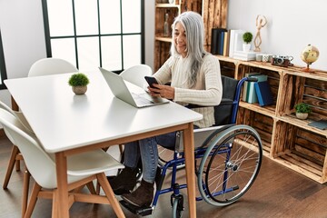 Middle age grey-haired woman using smartphone sitting on wheelchair at home
