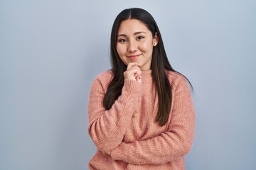 Young latin woman standing over blue background looking confident at the camera smiling with crossed arms and hand raised on chin. thinking positive.