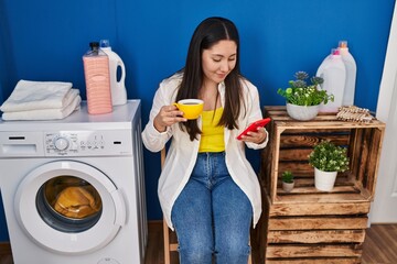 Young hispanic woman using smartphone and drinking coffee waiting for washing machine at laundry room