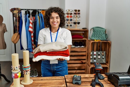 Young latin shopkeeper woman smiling happy holding stack of sweater at clothing store.