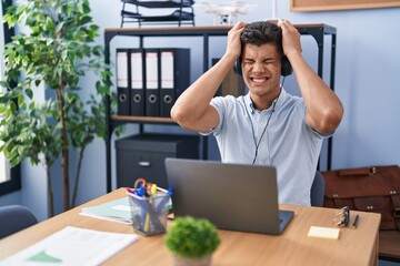 Young hispanic man working at the office wearing headphones suffering from headache desperate and...