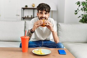 Young hispanic man eating classical burger and drinking soda sitting on the sofa at home.