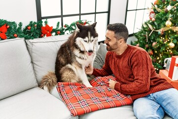 Young hispanic man smiling confident sitting on sofa with dog by christmas tree at home