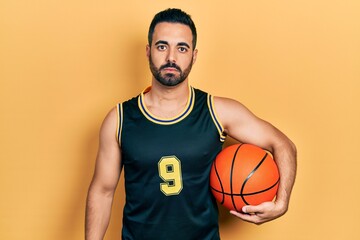 Handsome hispanic man with beard holding basketball ball thinking attitude and sober expression looking self confident