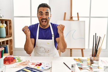 Young hispanic man with beard at art studio with painted face celebrating surprised and amazed for success with arms raised and open eyes. winner concept.