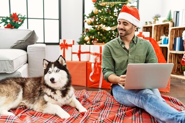 Obraz premium Young hispanic man using laptop sitting on floor with dog by christmas tree at home