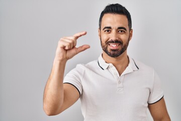 Young hispanic man with beard wearing casual clothes over white background smiling and confident gesturing with hand doing small size sign with fingers looking and the camera. measure concept.