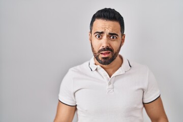 Young hispanic man with beard wearing casual clothes over white background in shock face, looking skeptical and sarcastic, surprised with open mouth