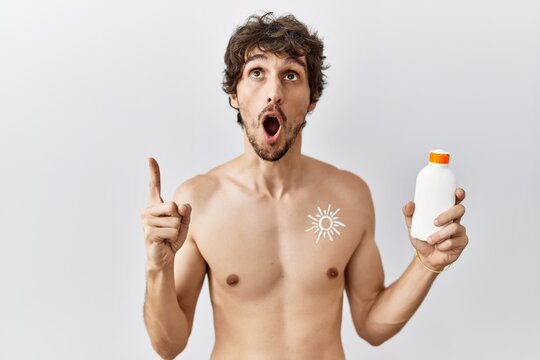 Young hispanic man standing shirtless holding sunscreen lotion amazed and surprised looking up and pointing with fingers and raised arms.