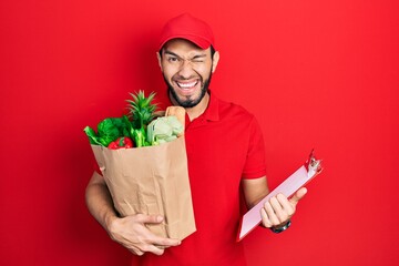 Hispanic man with beard wearing courier uniform with groceries from supermarket and clipboard...