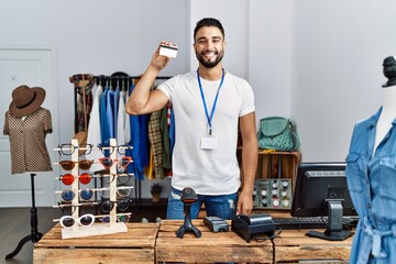 Young arab man shopkeeper holding credit card working at clothing store - obrazy, fototapety, plakaty