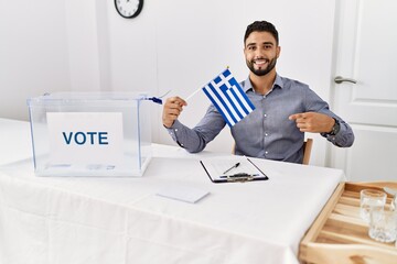 Young handsome man with beard at political campaign election holding greece flag smiling happy pointing with hand and finger