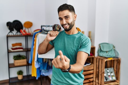 Young handsome man with beard holding shopping bags at retail shop beckoning come here gesture with hand inviting welcoming happy and smiling