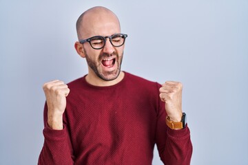 Young bald man with beard standing over white background wearing glasses celebrating surprised and amazed for success with arms raised and eyes closed. winner concept.