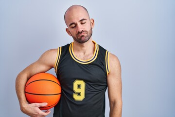 Young bald man with beard wearing basketball uniform holding ball looking sleepy and tired, exhausted for fatigue and hangover, lazy eyes in the morning.