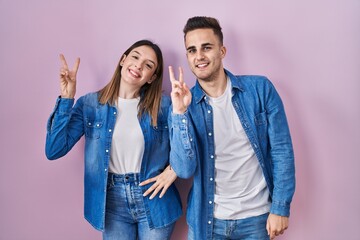 Young hispanic couple standing over pink background smiling looking to the camera showing fingers doing victory sign. number two.