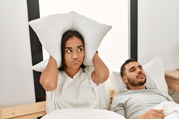 Woman covering ears with pillow irritated for snore lying in bed at home.