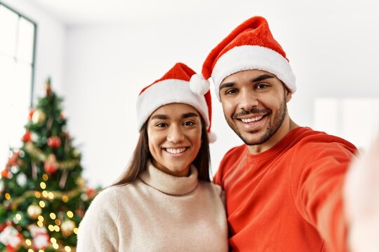 Young hispanic couple wearing christmas hat making selfie by the smartphone at home.
