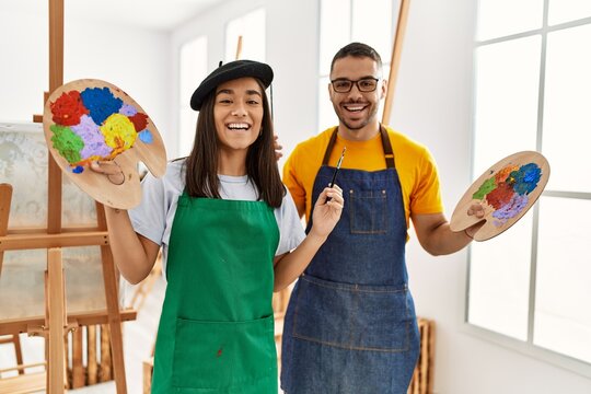 Young hispanic couple smiling happy holding paintbrush and palette at art studio.