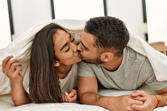 Young latin couple kissing and covering with bed sheet lying on the bed.