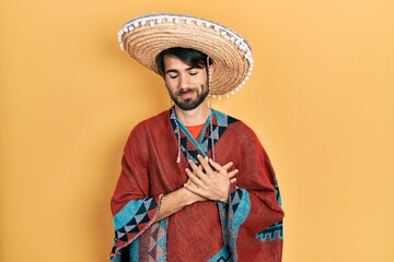 Young hispanic man holding mexican hat smiling with hands on chest with closed eyes and grateful gesture on face. health concept.