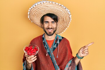 Young hispanic man wearing mexican hat holding chili smiling happy pointing with hand and finger to the side